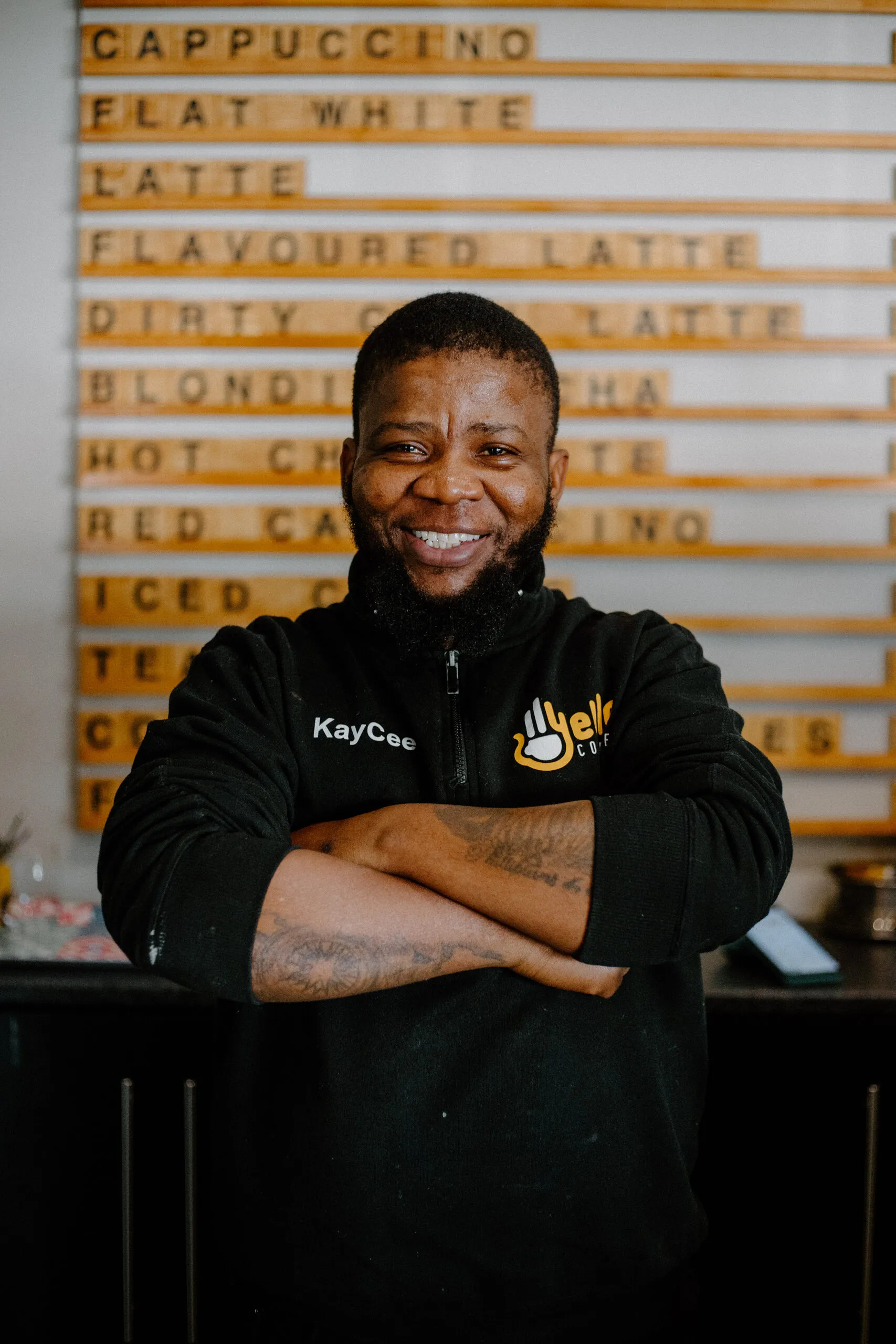 KayCee in a Yello Coffee jacket in front of the menu board